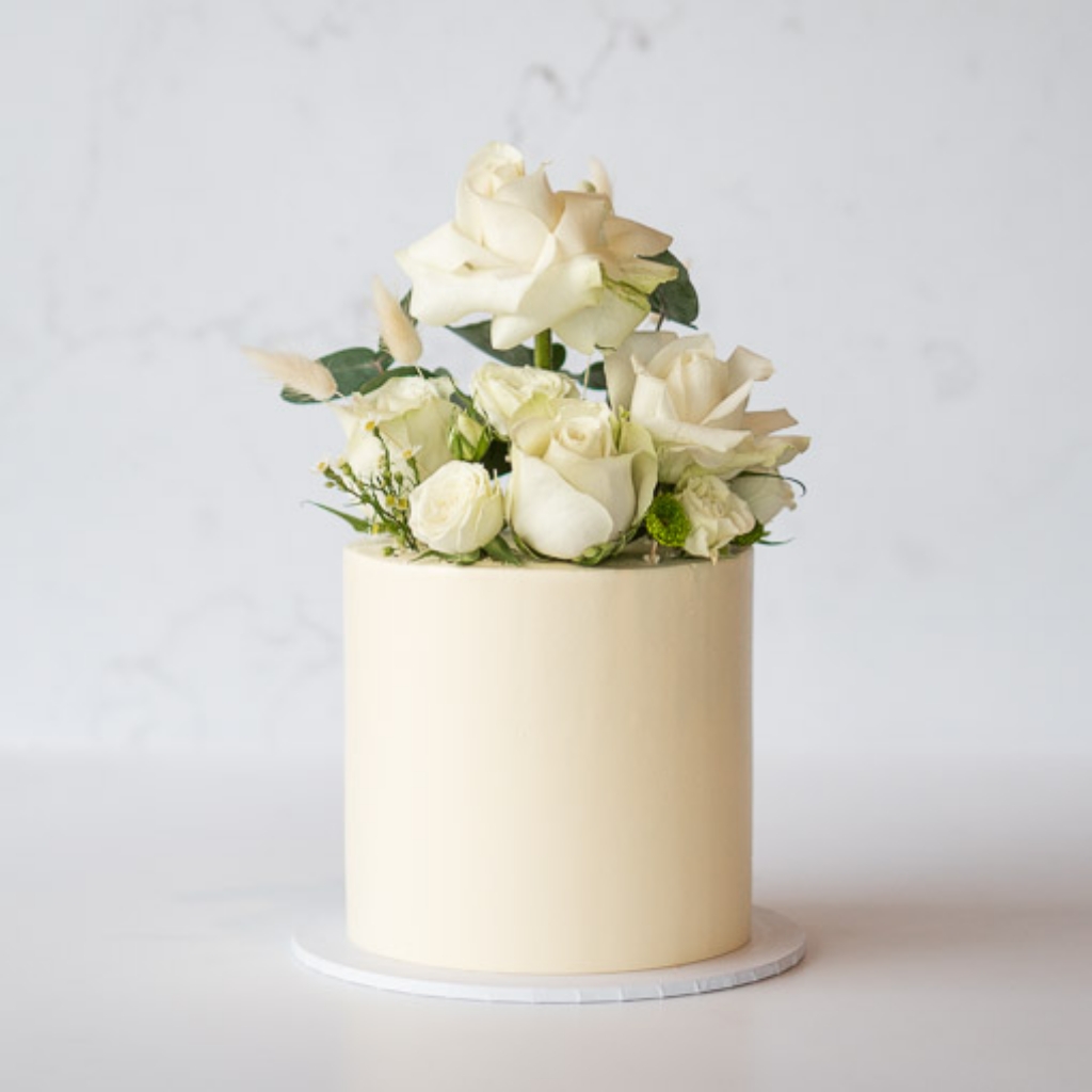  A smooth, light cream buttercream cake topped with a generous arrangement of fresh white roses and various green leaves.