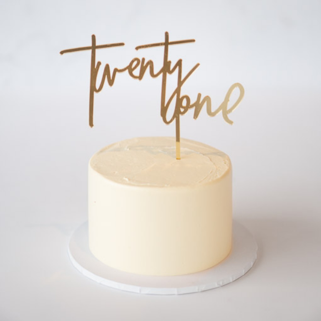 A gold-colored cake topper spelling "twenty one" in a large, elegant script font, placed vertically on top of a plain cream-colored round buttercream cake.