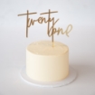 A gold-colored cake topper spelling "twenty one" in a large, elegant script font, placed vertically on top of a plain cream-colored round buttercream cake.