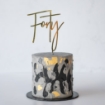 A gold-colored cake topper spelling "forty" in a large, elegant script font, placed vertically on top of a plain cream-colored round buttercream cake.