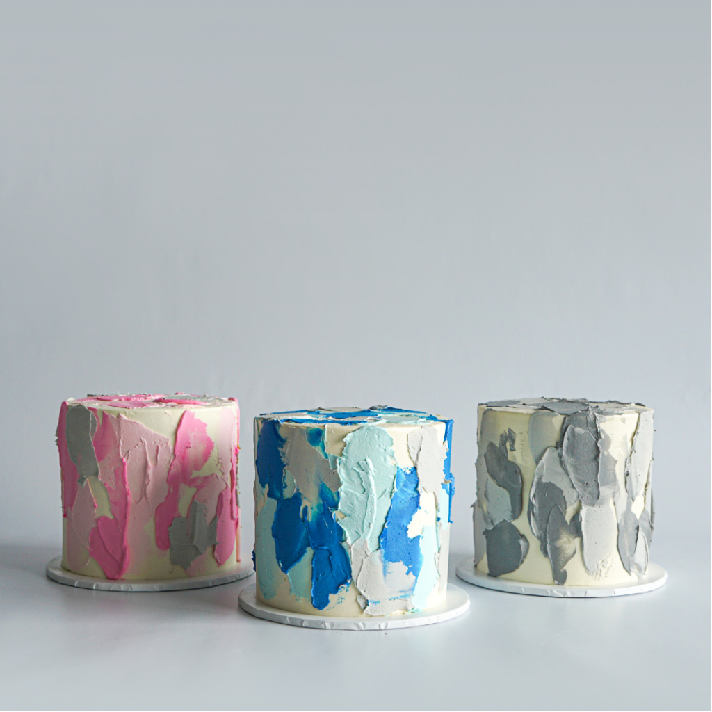 Three modern buttercream cakes featuring a rough, abstract watercolour painted texture. The cake on the left is pink, the middle cake is blue, and the cake on the right is grey.