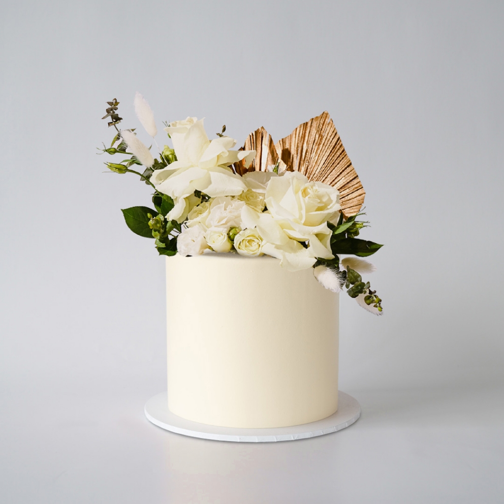 A pristine white or light cream buttercream cake, adorned on top with a large gold fan-shaped decoration, a cluster of fresh white roses, and various green leaves and dried white floral accents.