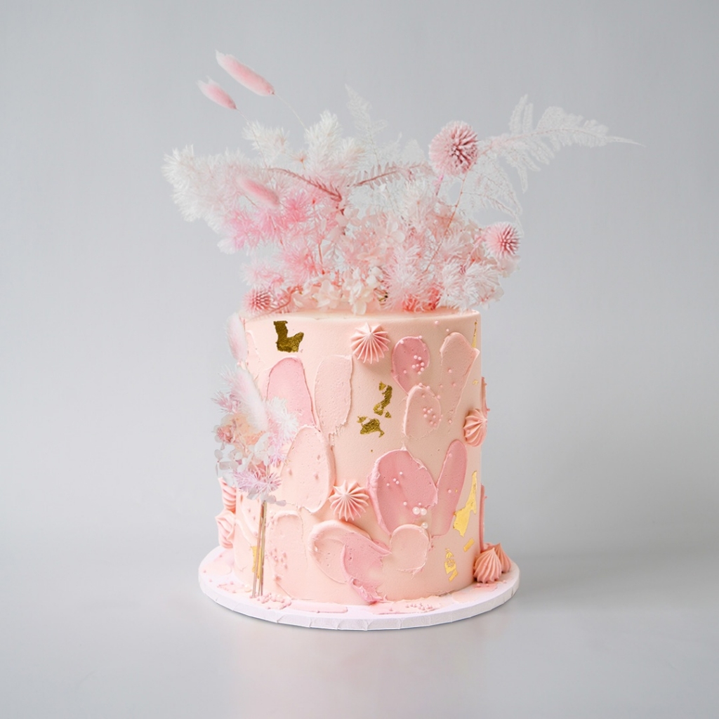 A light pink buttercream cake with textured pink painted strokes, gold foil accents, pink meringue kisses, and a dramatic top arrangement of pink and white dried florals and plumes.