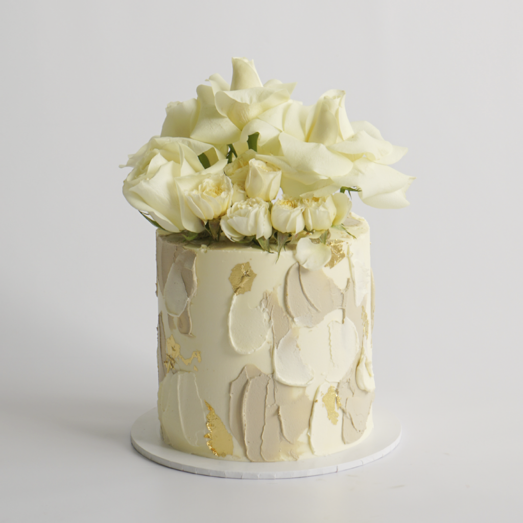 A round buttercream cake with a rough, abstract painted texture in shades of cream and light beige, topped with a cluster of white roses.
