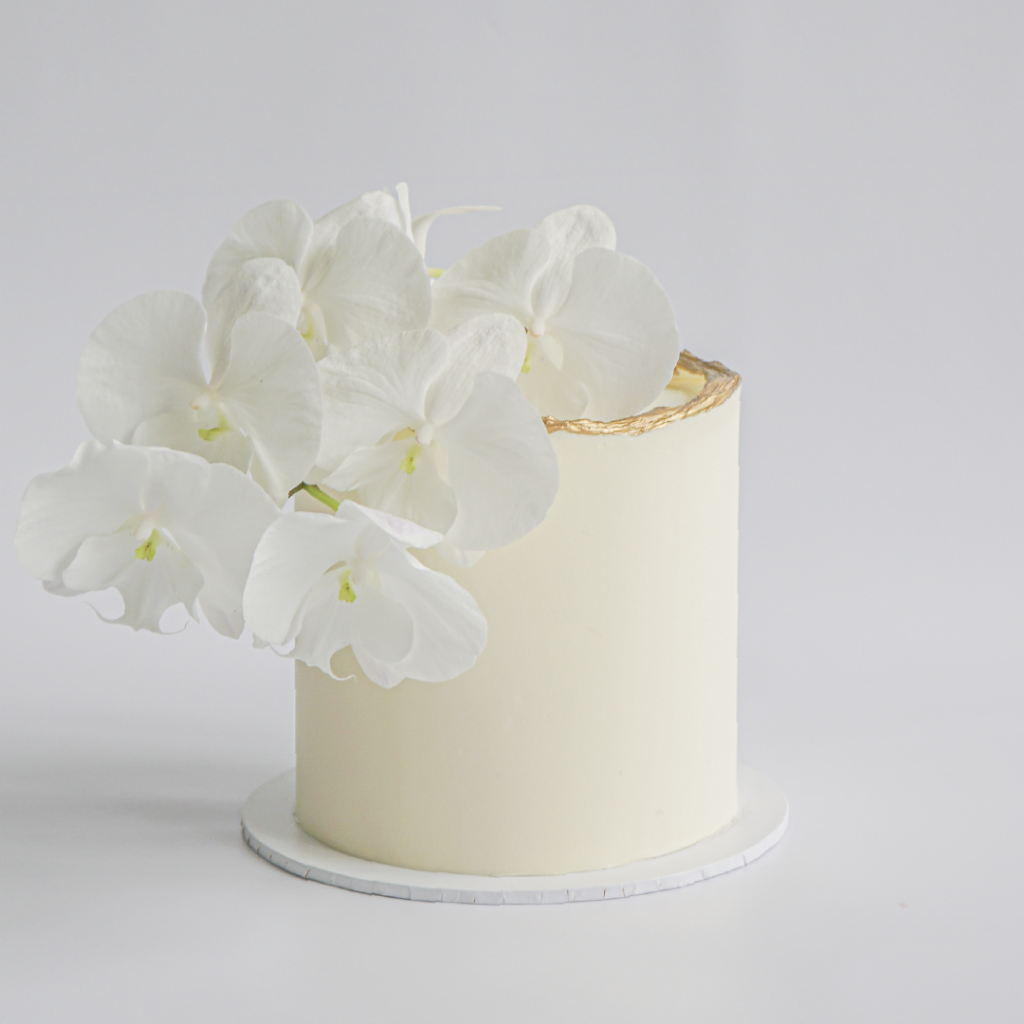 A pristine cream-colored buttercream cake with a textured gold border along the top edge, lavishly decorated with a cluster of white phalaenopsis orchids cascading down one side.