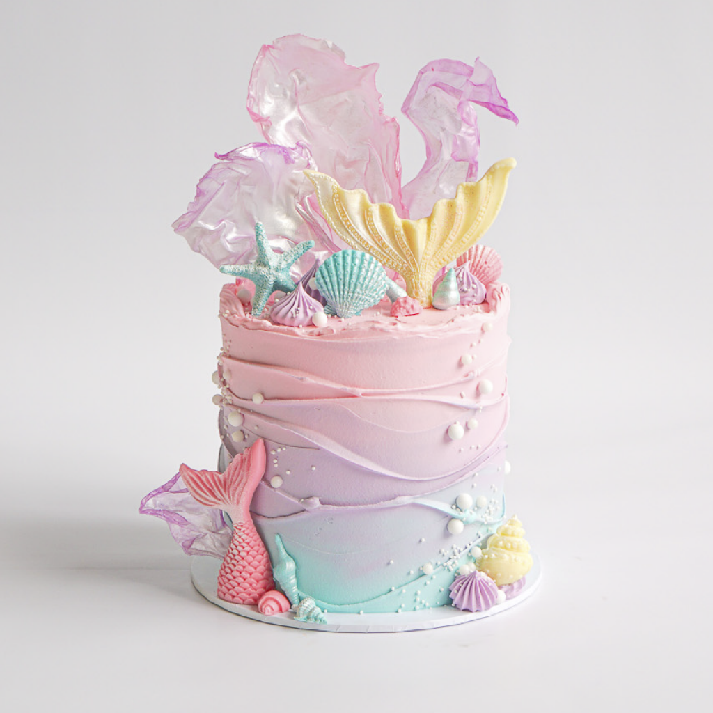 A round buttercream cake with ombre frosting blending from pink at the top, to purple in the middle, to light blue at the bottom, mimicking an underwater scene. The cake is decorated with white edible pearls, various edible seashells, and a pink mermaid tail fin emerging from the bottom. On top, there's a large yellow mermaid tail fin, a blue starfish, and translucent pink and purple sugar paper resembling ocean waves or coral.
