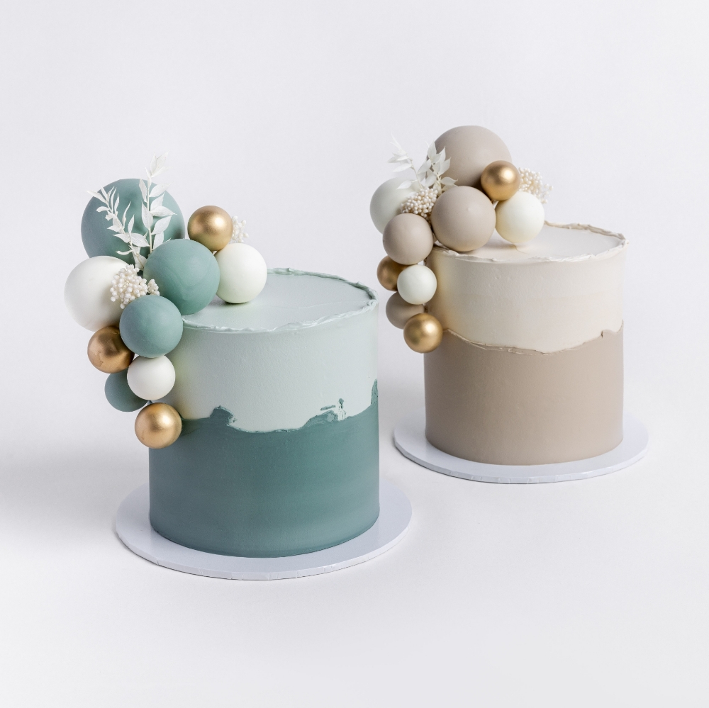Two modern buttercream cakes side by side, both with two-tone frosting and decorated with clusters of matte and metallic spheres.