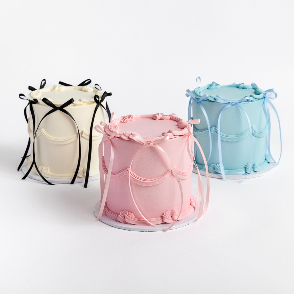 Three Buttercream Bow Cakes in white with black bows, pink with pink bows, and blue with blue bows, featuring cascading ribbons.
