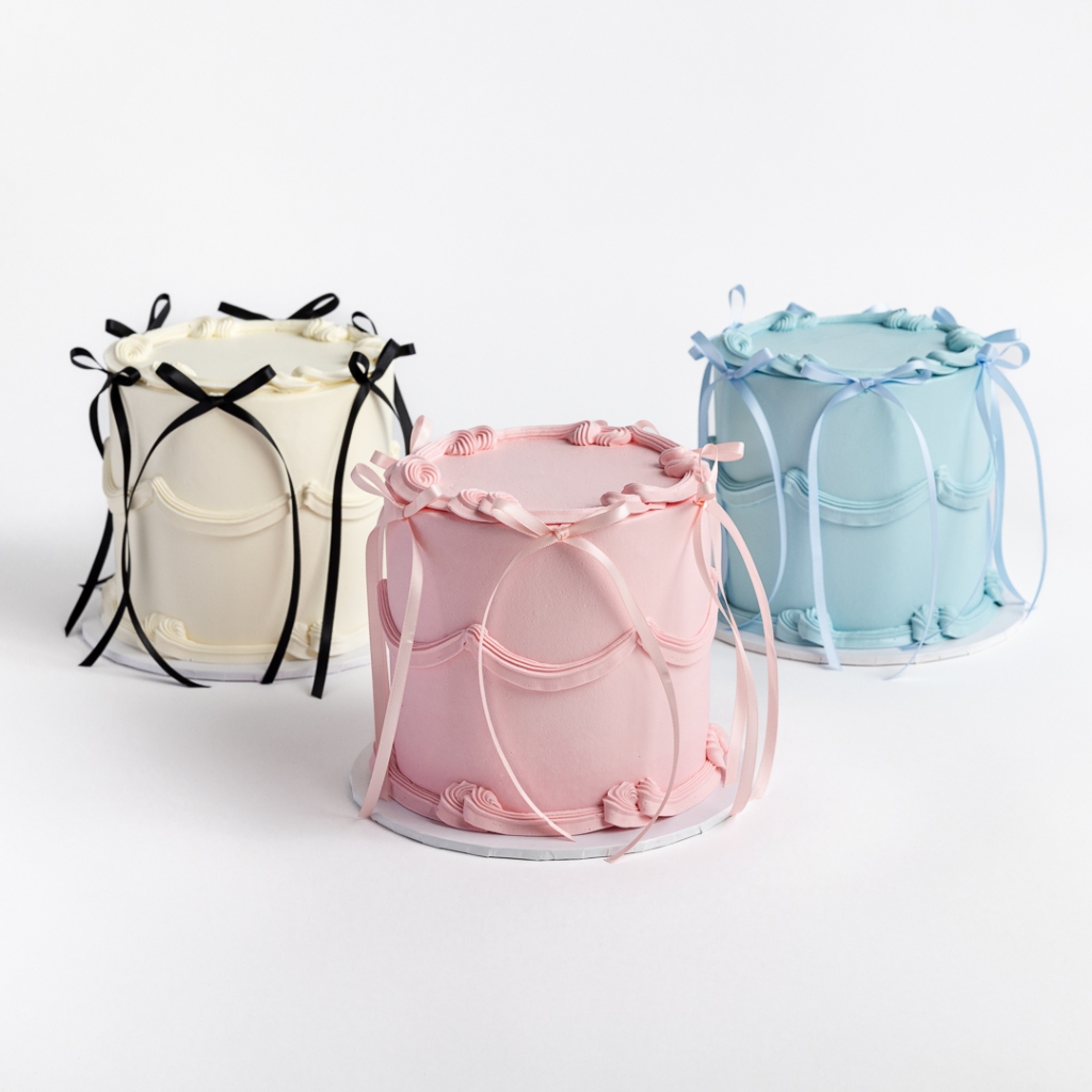 Three Buttercream Bow Cakes in white with black bows, pink with pink bows, and blue with blue bows, featuring cascading ribbons.
