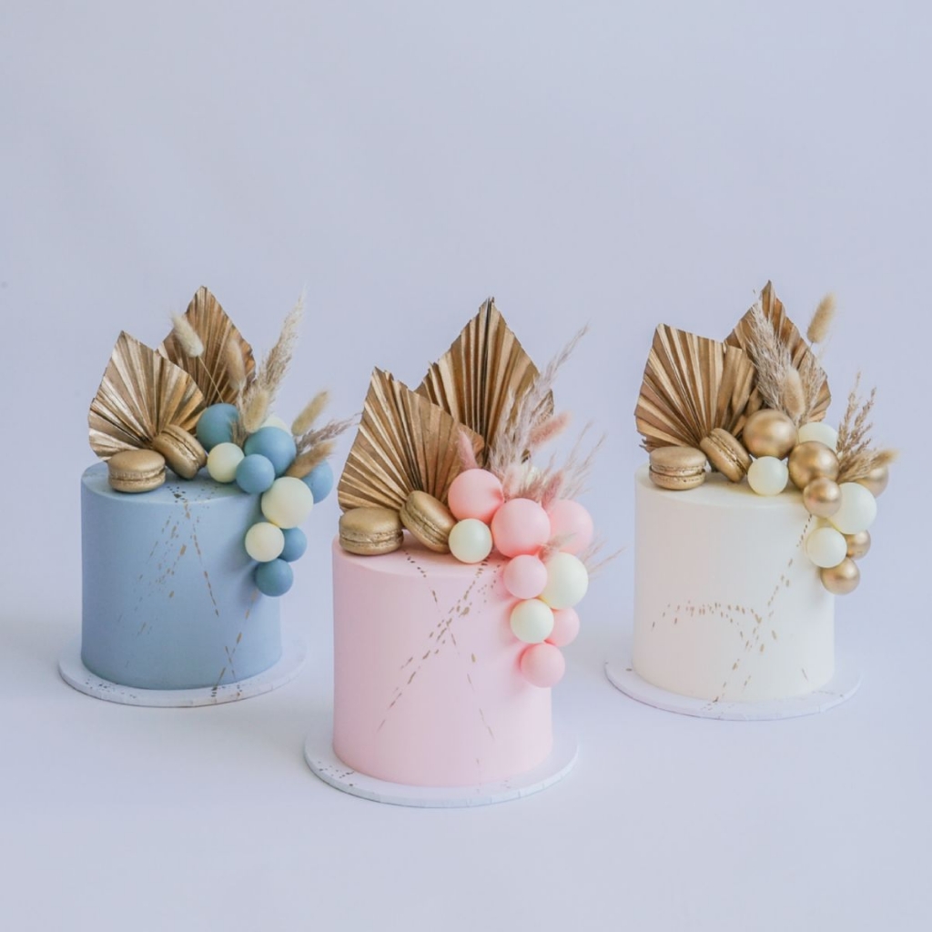 A trio of modern buttercream cakes in pastel blue, pink, and cream, each topped with a stunning fancy faux floral arrangement of gilded palm spears and macarons.
